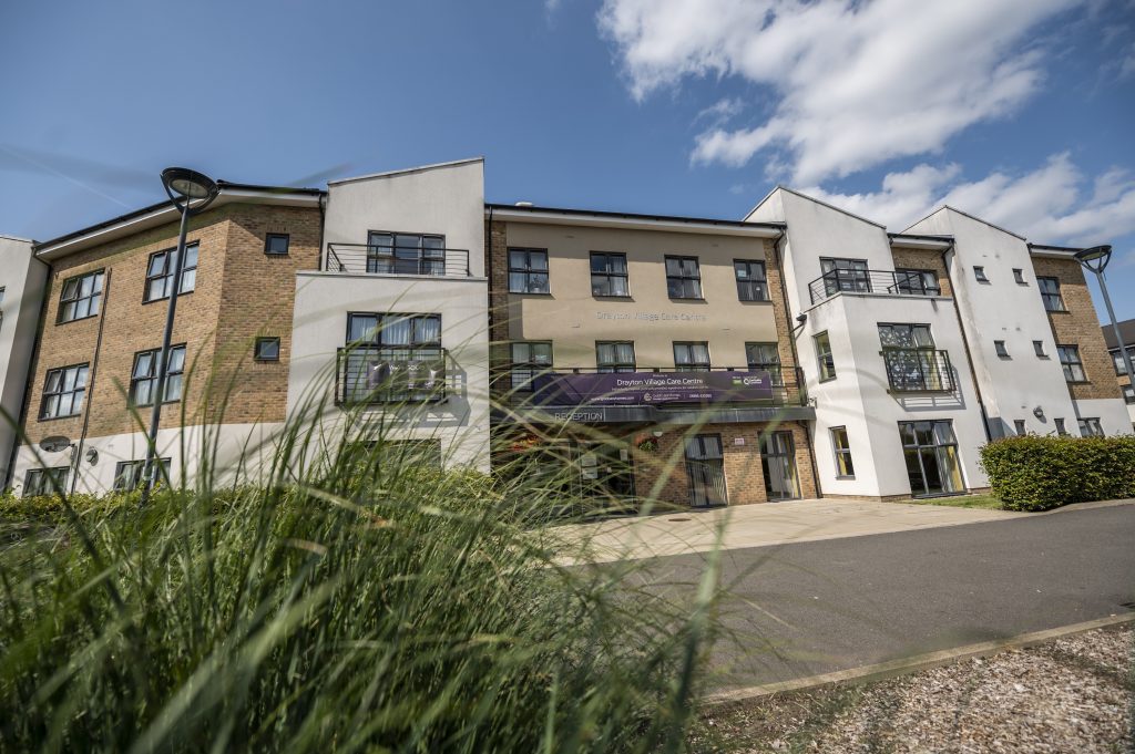 Modern care home building with a mix of brick and white exterior, featuring balconies, greenery in the foreground, and a clear blue sky.