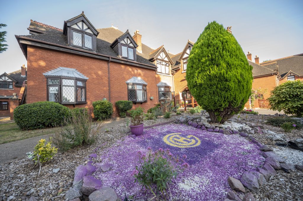 Welcoming garden at Heath Lodge featuring a vibrant purple flower bed, a manicured tree, and a charming red-brick building.