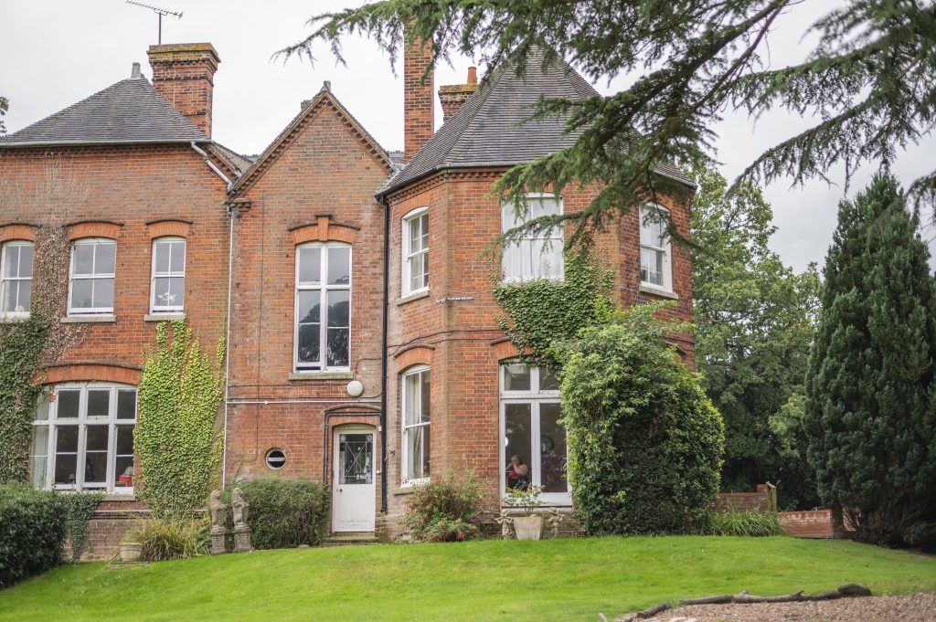 Victorian-style care home with red brick exterior, large windows, and greenery, set in a manicured garden.