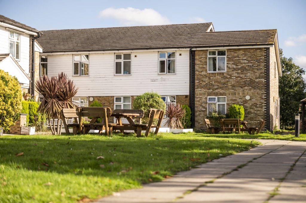 Two-story nursing home building with a mix of brick and white siding, surrounded by grassy areas and wooden benches under clear blue skies.