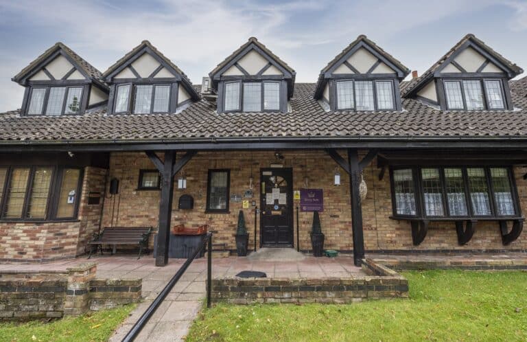 Traditional Tudor-style care home exterior with a brick façade, pitched roof, and multiple gabled windows, set in a grassy area.