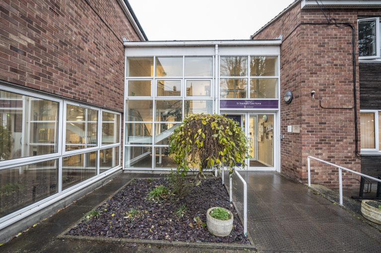 Modern brick building entrance featuring large glass panels, a small landscaped area with a bush, and signage for St Stephen's Care Home.