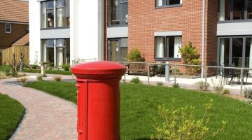 Bright red post box near a landscaped path leading to a modern care home with a mix of brick and white exterior. Green grass and shrubs surround the area, Amy Woodgate House, a care home in Chessington.