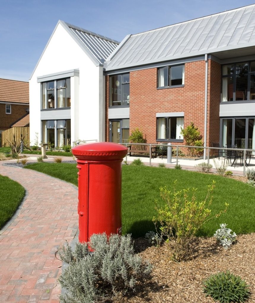 Bright red post box near a landscaped path leading to a modern care home with a mix of brick and white exterior. Green grass and shrubs surround the area.
