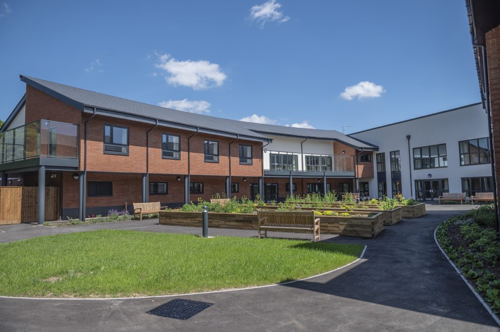 Modern two-story nursing home building with red brick and glass balconies, surrounded by landscaped gardens and benches under a blue sky.