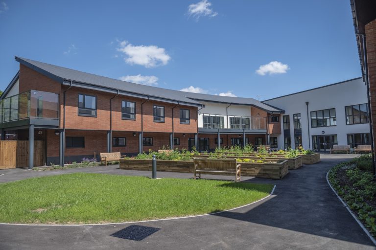 Modern two-story nursing home building with red brick and glass balconies, surrounded by landscaped gardens and benches under a blue sky.