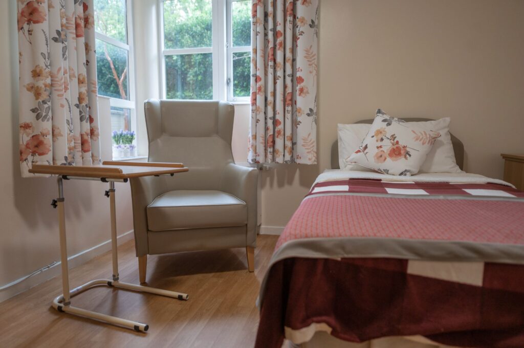 A cozy bedroom featuring a single bed with a red and white quilt, a floral cushion, a gray armchair, and light-colored curtains.