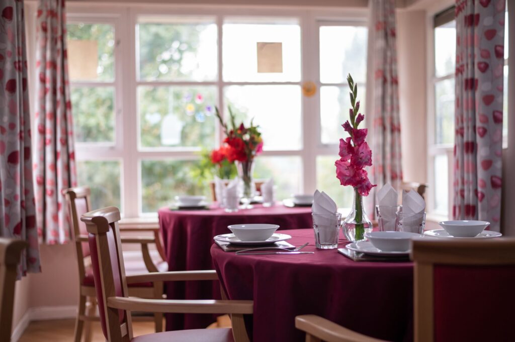 A dining area with tables set in red tablecloths, featuring flower centerpieces and large windows letting in natural light.