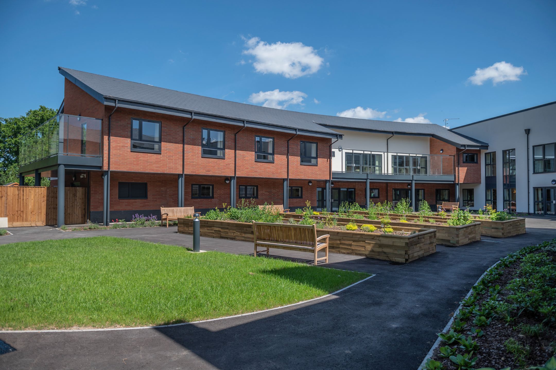 Modern nursing home building with brick and glass facade, surrounded by landscaped gardens and vibrant planters under a blue sky.