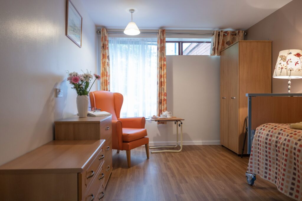 Bright, cozy room featuring an orange armchair, wooden dresser, and bedside table, with natural light filtering through sheer curtains.