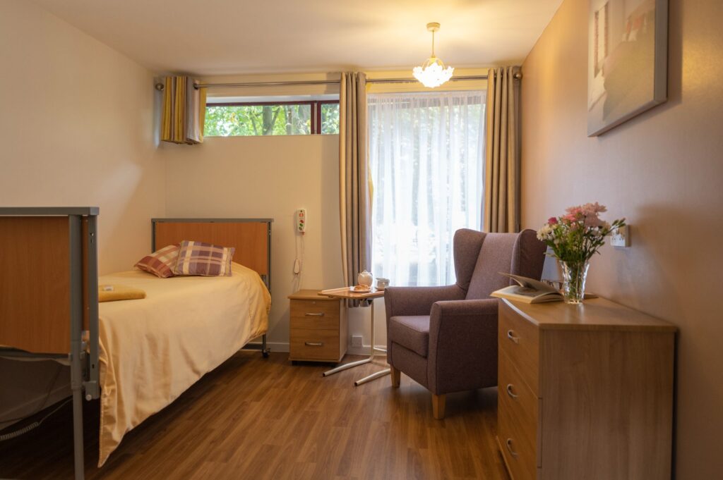 Cozy care home room featuring a single bed, armchair, bedside table, and a window with sheer curtains, accentuated by warm tones.