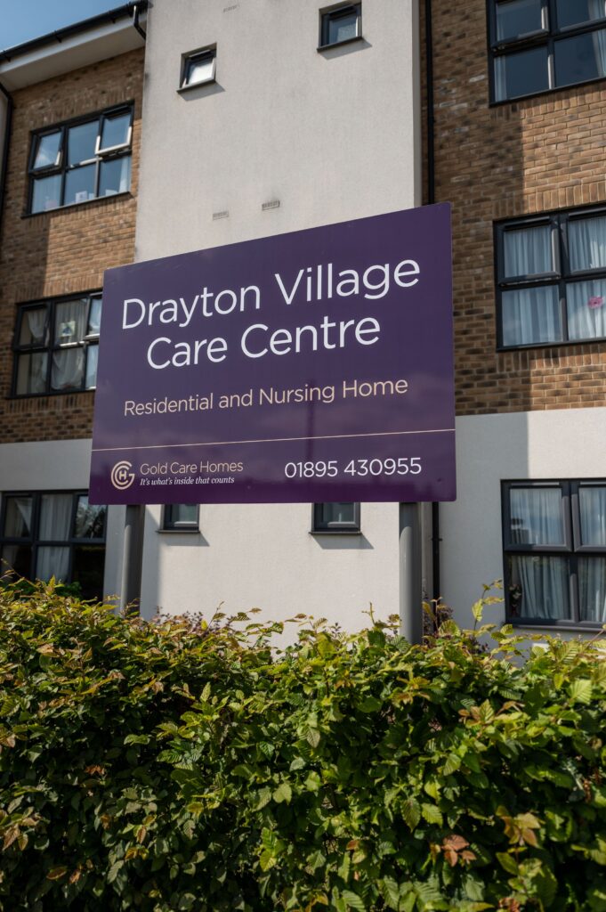 Drayton Village Care Centre sign in front of the care home's building with windows and greenery in the foreground.
