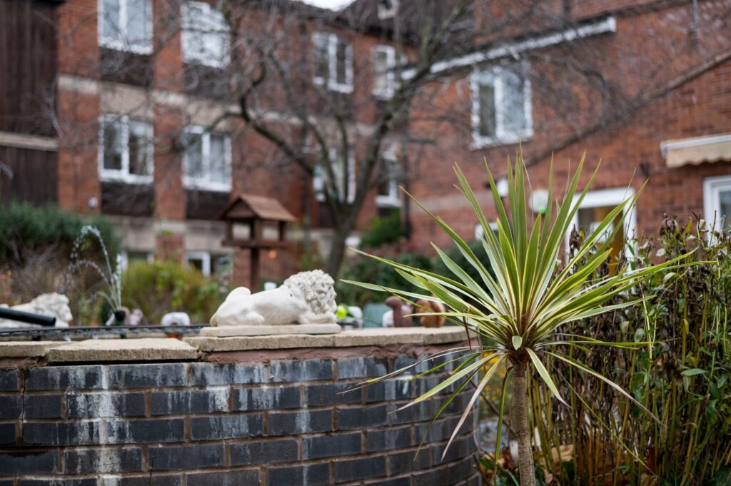 A landscaped garden at St. Stephen's Care Home features a stone wall, a lion statue, and a spiky green plant, with a backdrop of brick buildings.