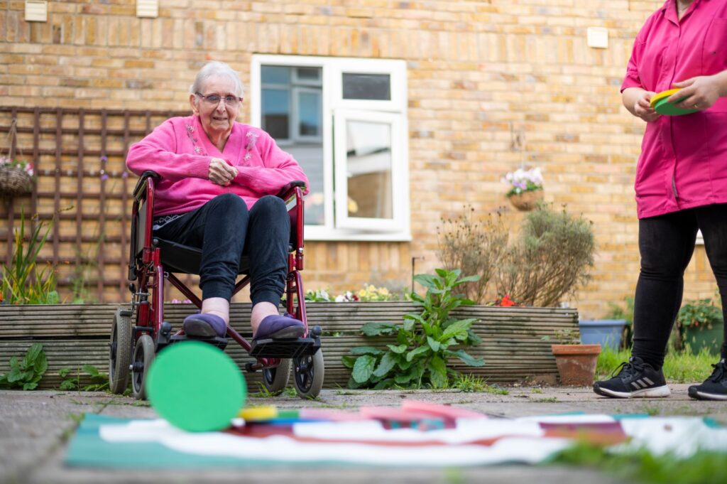 Elderly woman in a wheelchair smiles in a garden while a caregiver holds colorful shapes, surrounded by greenery and flowers.