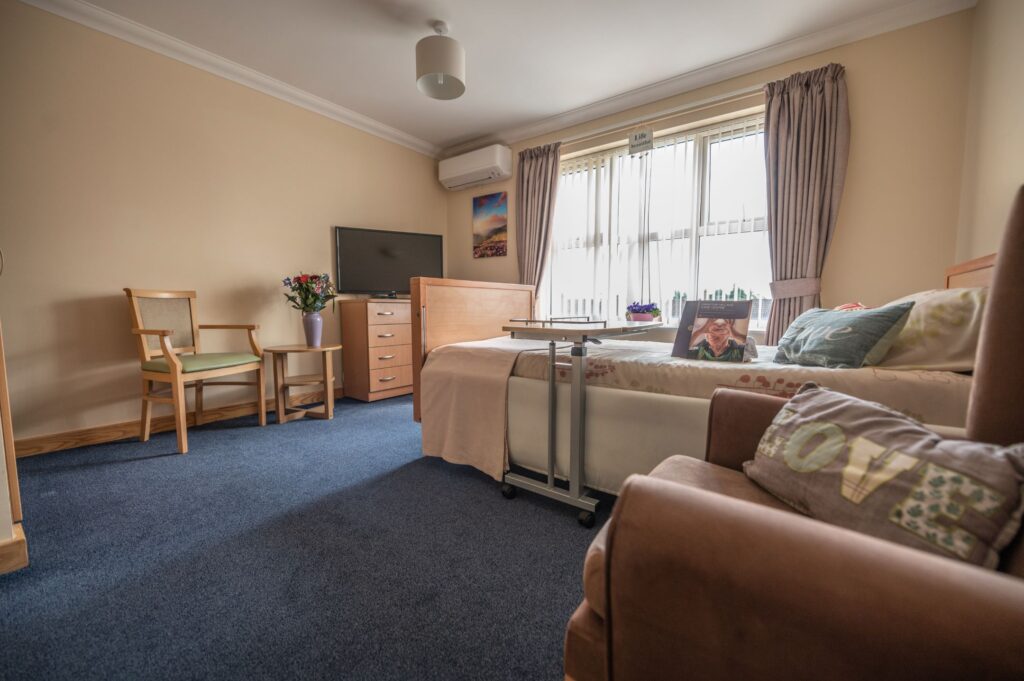 Comfortable and well-lit bedroom in a care home in West Drayton, featuring a bed, armchair, TV, and flowers on a side table.
