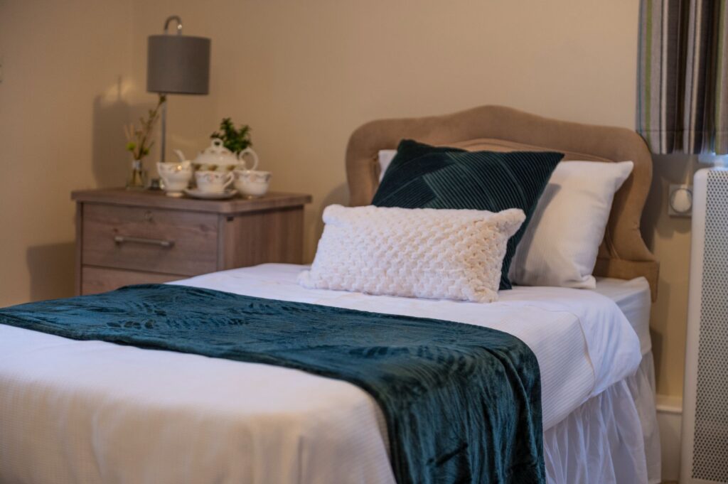 Cozy bedroom featuring a neatly made bed with a teal throw, decorative cushion, wooden nightstand, and teapot set.