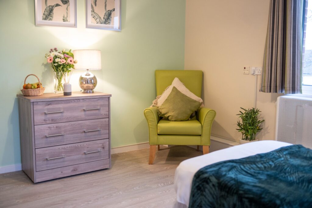 Cozy room featuring a green armchair, bedside table with flowers and lamp, and a light wood dresser. Soft colors create a welcoming atmosphere.