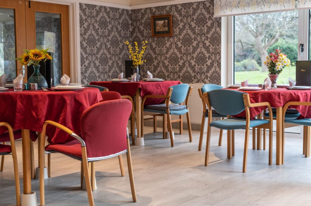 Dining room with red tablecloths, floral centrepieces, and a mix of blue and red chairs, set near a large window.