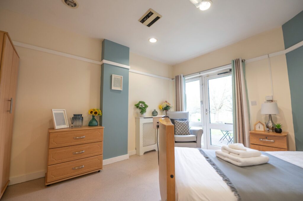 Spacious care home in Wantage bedroom with a neatly made bed, wooden furniture, and seating area by garden-facing doors.