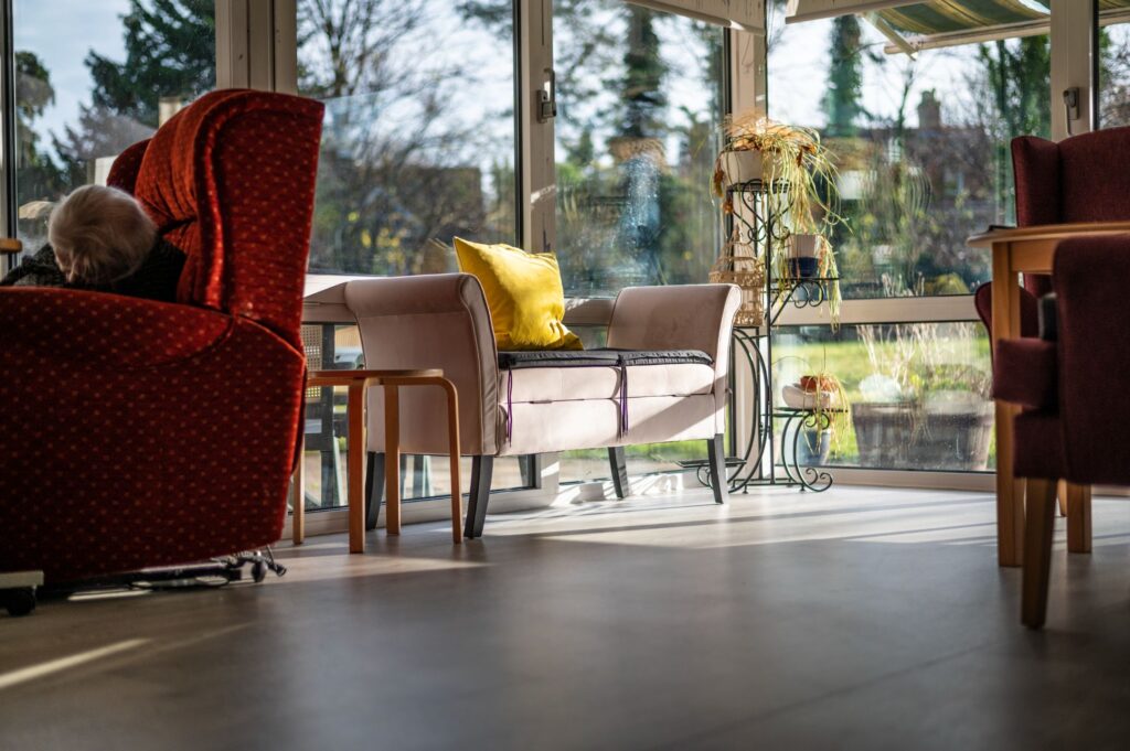 Sunlit care home lounge with a mix of red and beige seating, a yellow cushion, and large windows overlooking the garden.