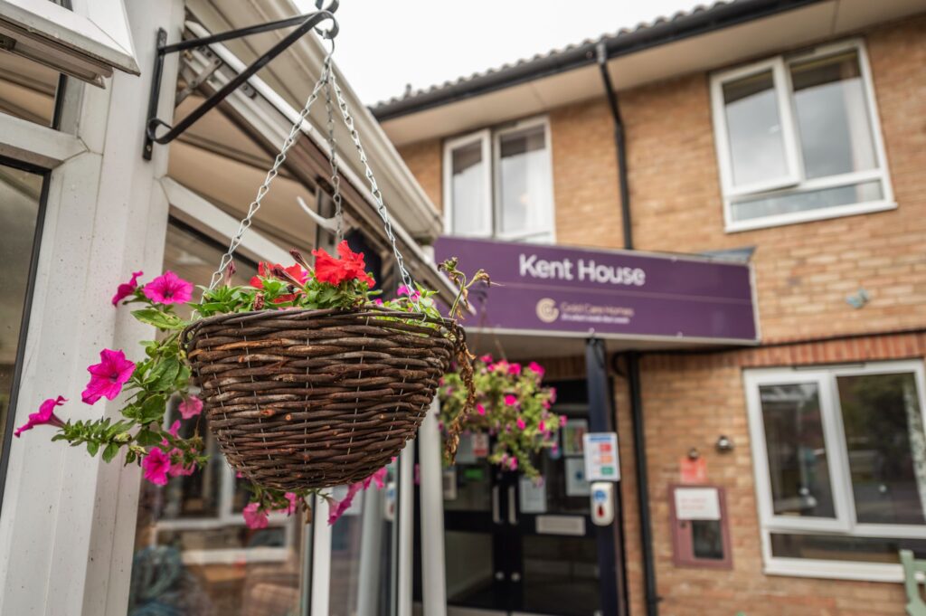 Hanging flower basket with pink and red blooms near the entrance of Kent House, showcasing a welcoming facade and signage.