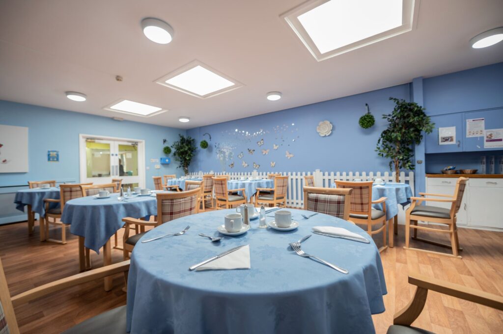Bright dining area with blue walls and skylights, featuring round tables with blue tablecloths, neatly set for tea with cups and saucers.