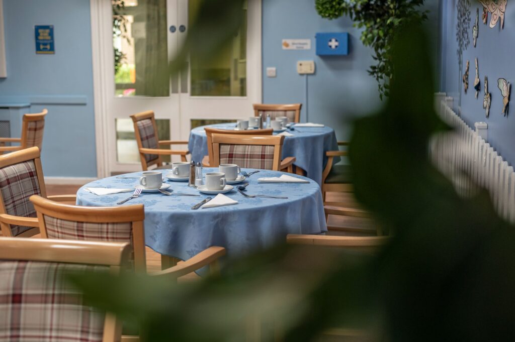 Dining area in a care home, with round tables covered in blue tablecloths, set for tea with cups and saucers, surrounded by wooden chairs with plaid cushions.