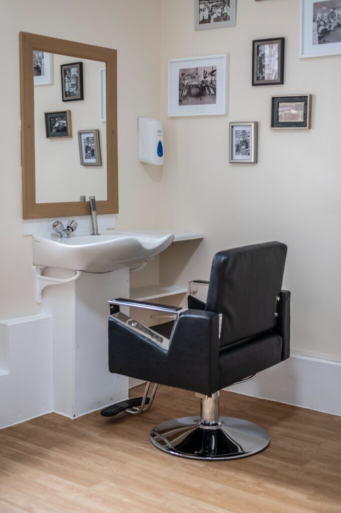 Hairdressing station in a care home, featuring a black salon chair, a mirror, and framed black-and-white photographs on the wall.