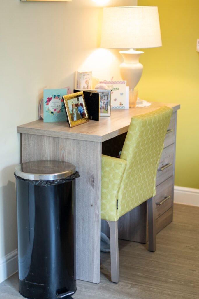 A modern desk with a yellow patterned chair, a lamp, photo frames, and a black trash can against a light green wall.