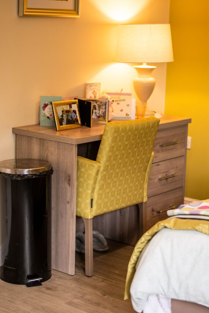 Cozy room corner featuring a wooden desk with a yellow upholstered chair, framed photos, a lamp, and a black trash bin.
