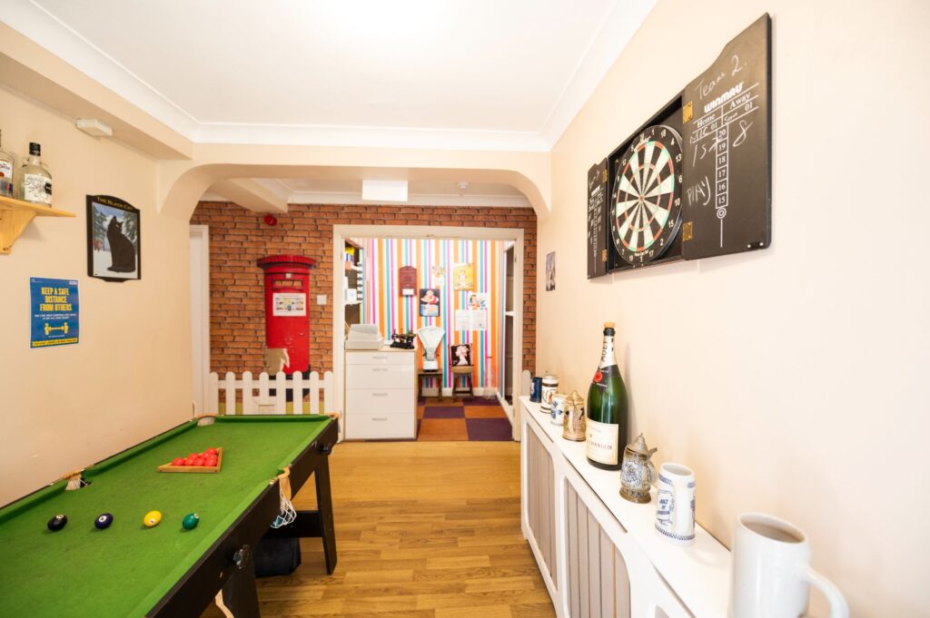 A bright communal area featuring a pool table, dartboard, and a cozy reception space with colorful decorations and a classic red post box.