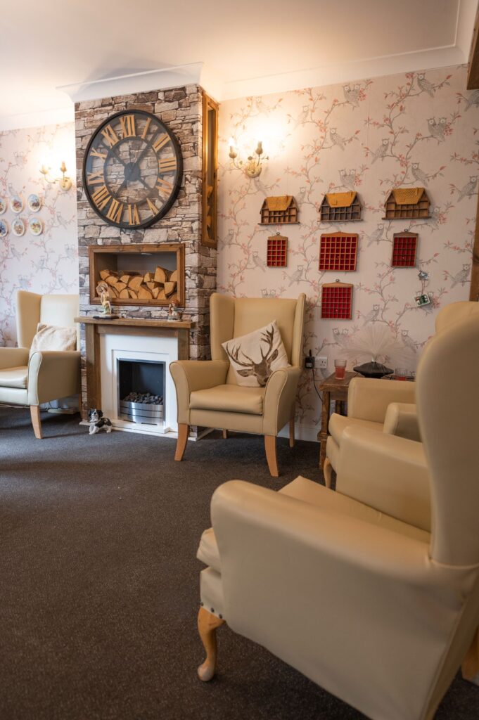Cozy lounge area featuring cream armchairs, a stone fireplace, a large clock, and decorative wall elements in soft colors.