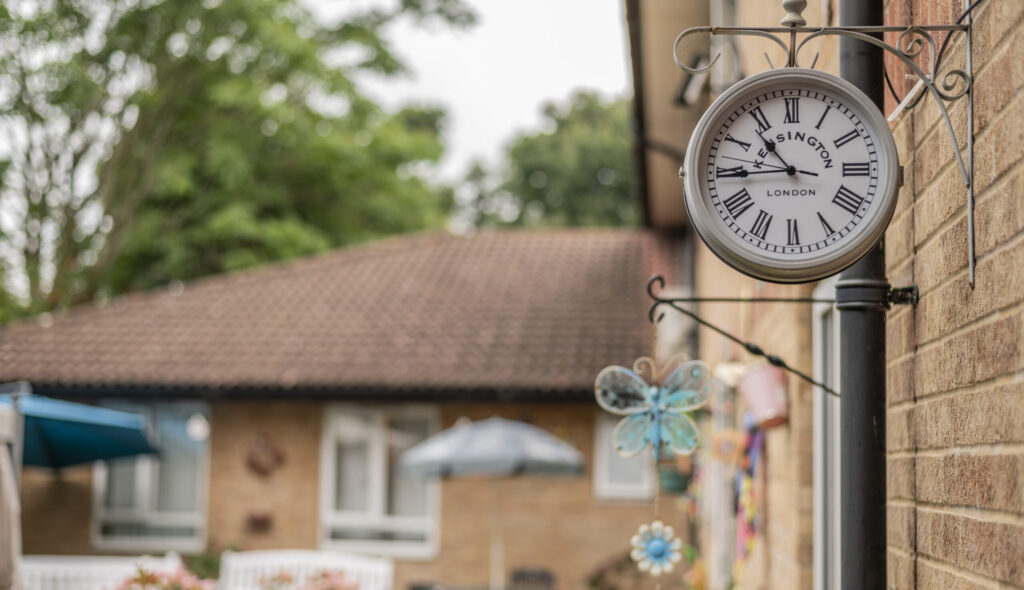 Traditional clock mounted on a wall, with a garden view featuring an umbrella and decorative flowers in the background.