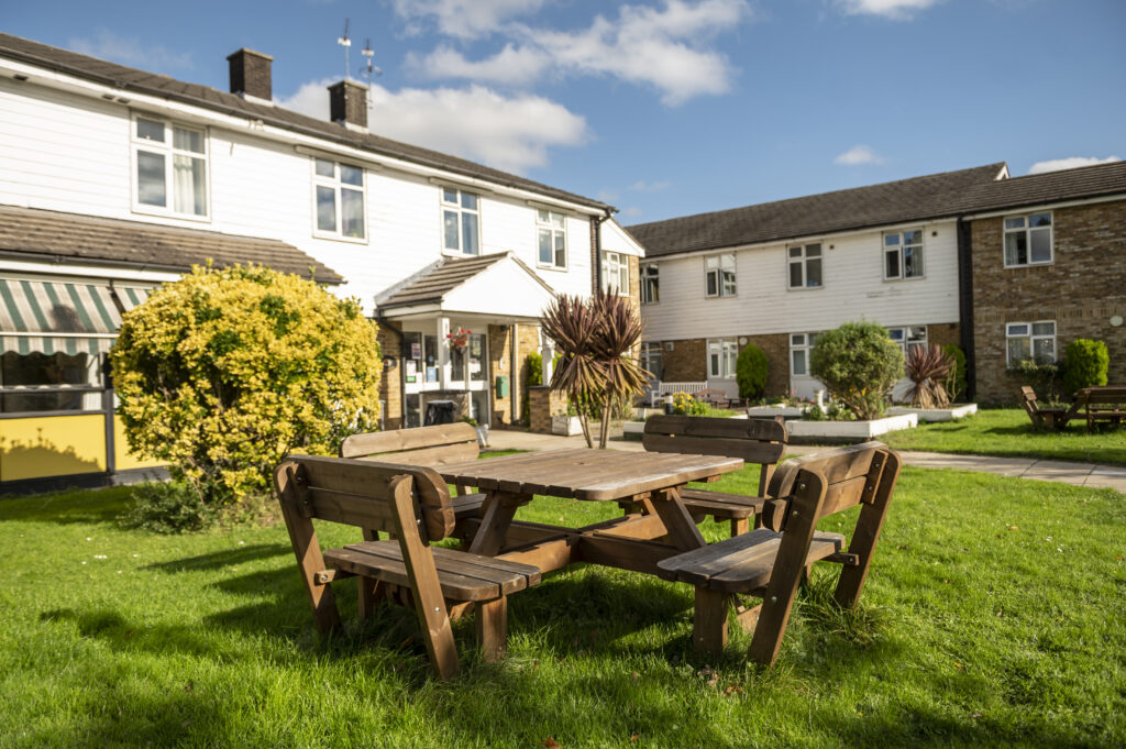 Wooden picnic table surrounded by chairs in a green lawn, with two buildings in the background and blue skies overhead at Hillside Nursing Home.