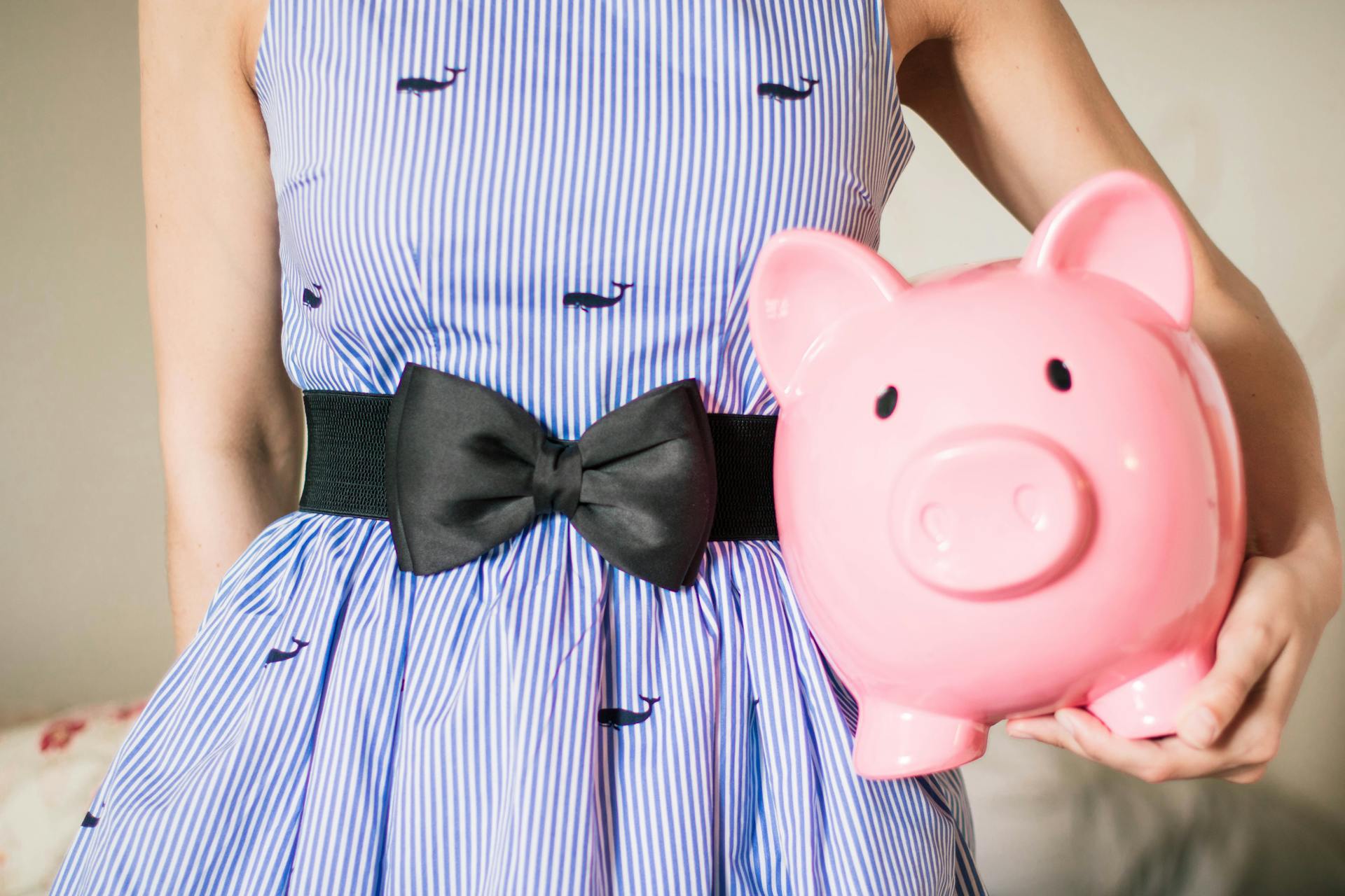 A woman in a blue striped dress with a black bow holds a pink piggy bank, emphasizing a cheerful savings theme.