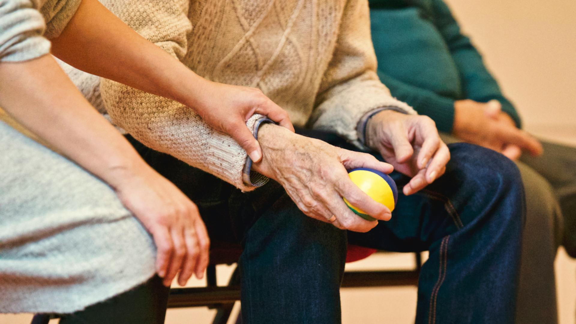A tender moment in a care home setting where a younger woman, dressed in a soft jumper, is gently holding the hands of an elderly man. The man, who is seated and wearing a jumper and jeans, is gripping a small yellow ball with both hands, likely as part of a therapeutic exercise. They are seated close together, symbolising care and support.