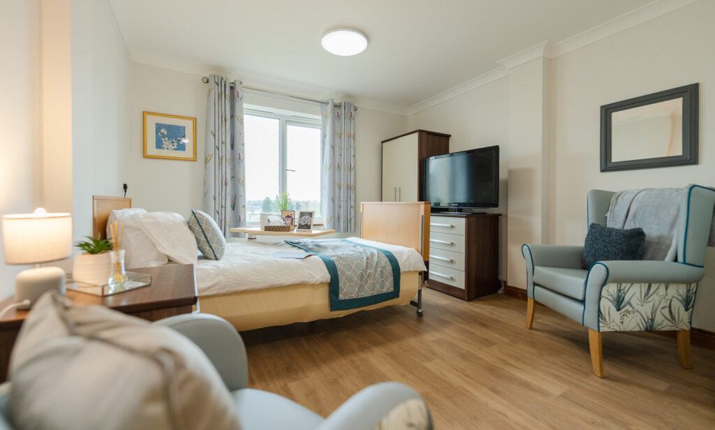 Bright and cosy care home bedroom with a comfortable bed, armchair, and large TV, featuring personal touches and soft lighting.