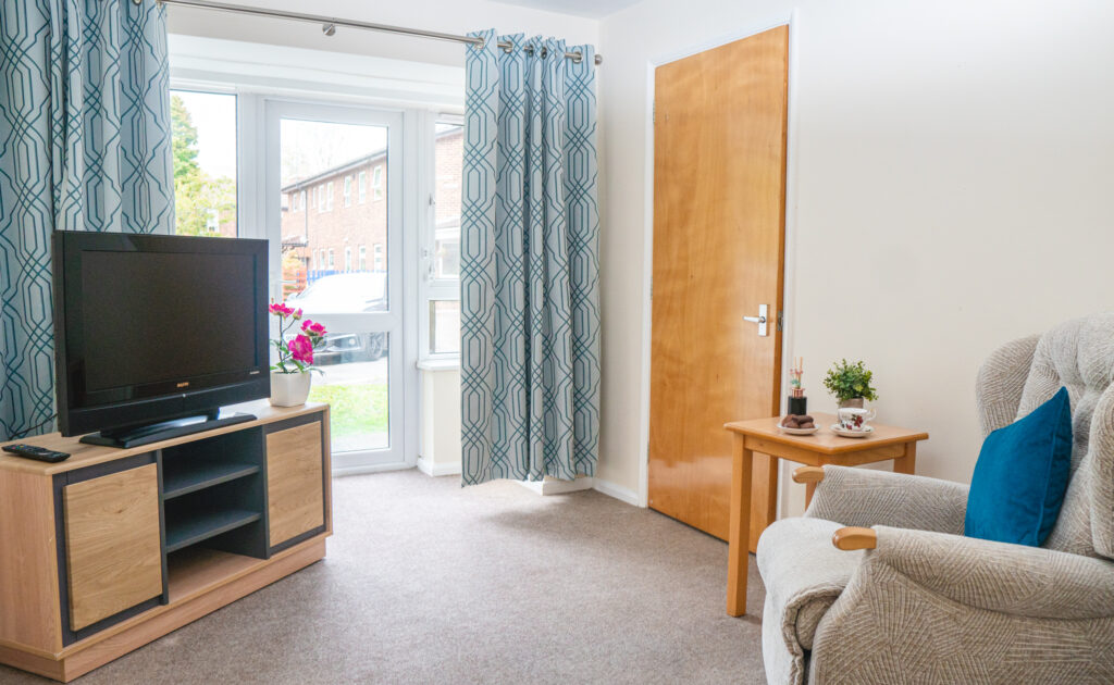 Comfortable living room in a care home with a TV on a wooden stand, a cosy armchair, and a side table with tea and snacks by the window.