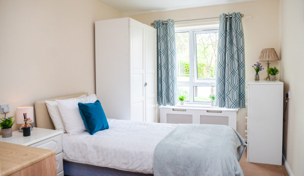 Bright and cosy bedroom in a care home in Lucton, featuring a neatly made bed with blue and white decor, a wardrobe, and a window with patterned curtains.