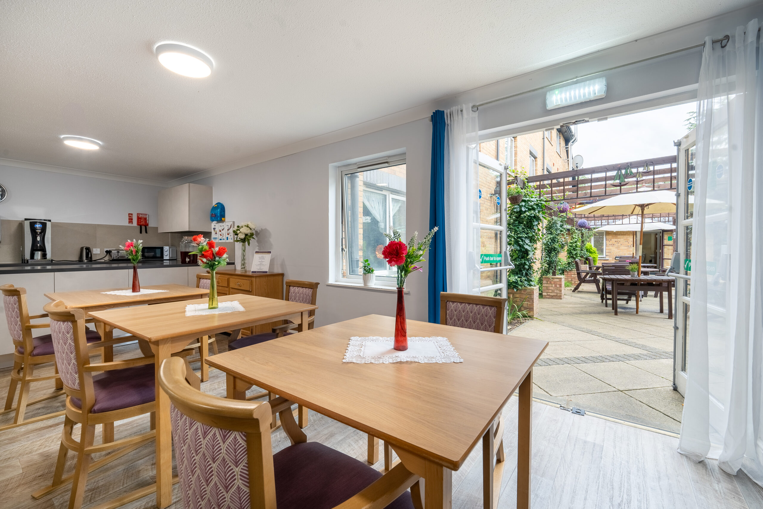 Dining area at Norwood Green Care Home, with wooden tables, chairs, and vases of flowers, leading to an outdoor patio with tables and umbrellas.
