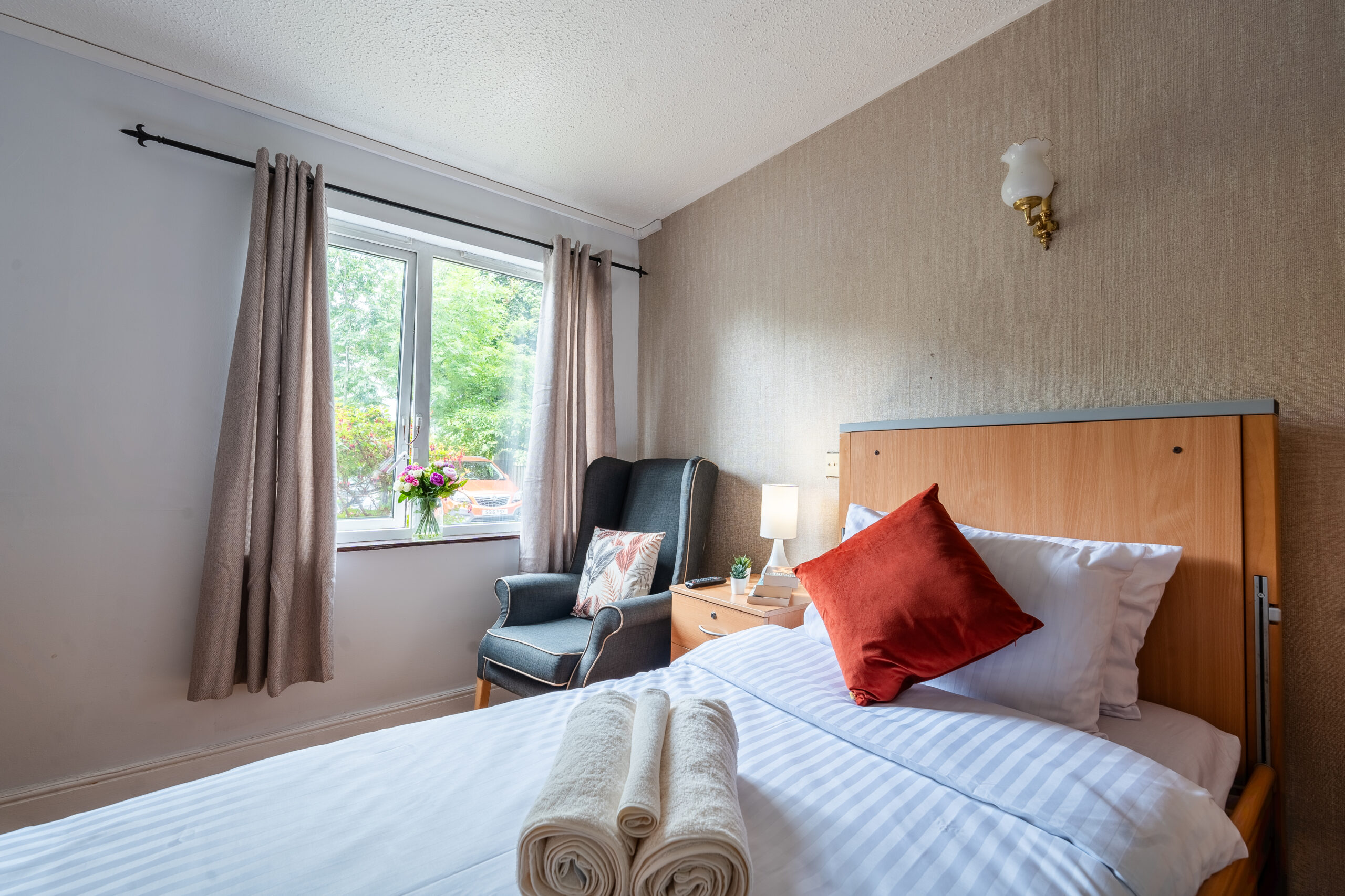 Comfortable bedroom in a care home in Southall, featuring a neatly made bed with orange accents, an armchair by the window, and natural light coming through the curtains.