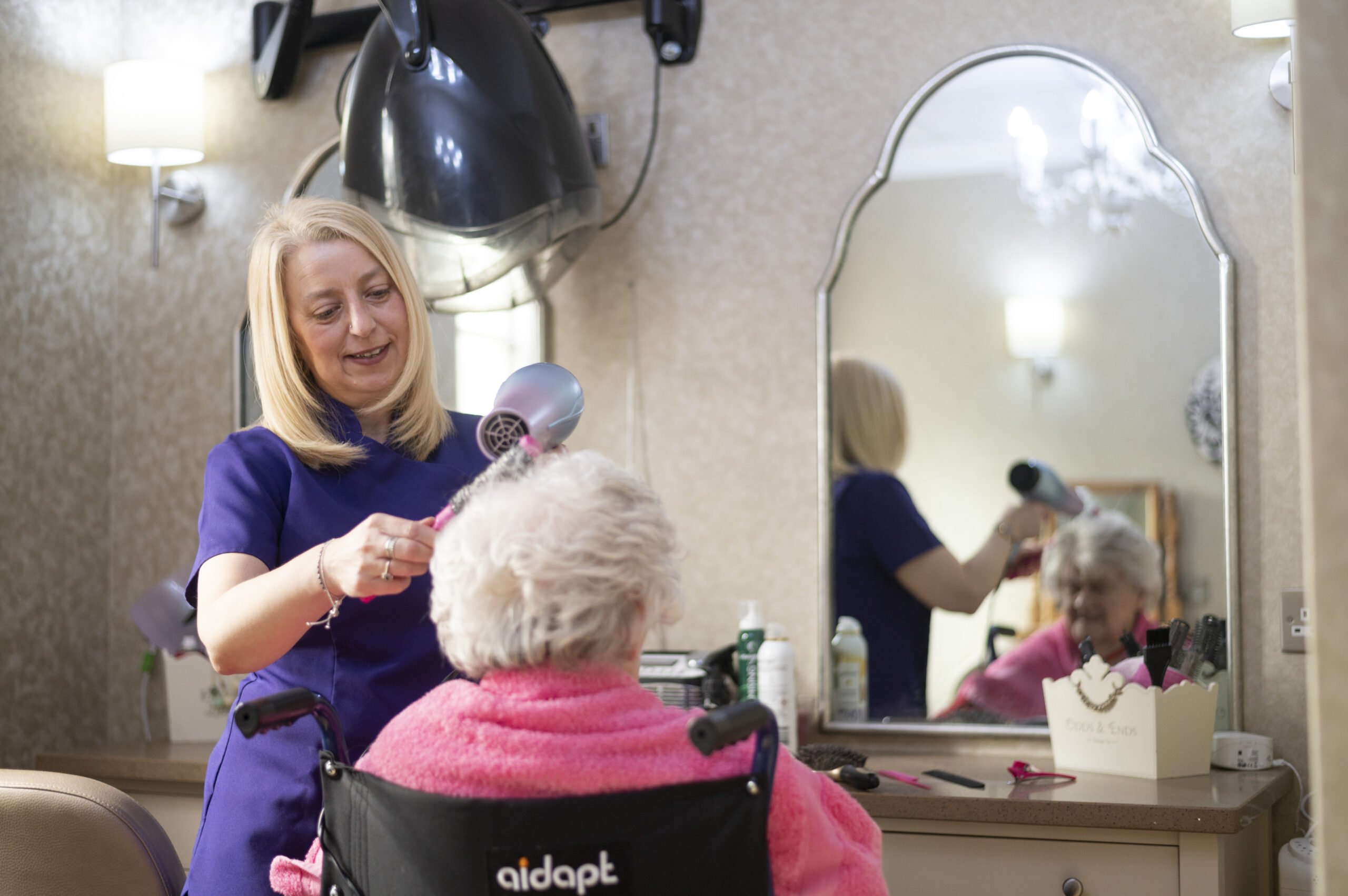 Care home staff member styling an elderly resident's hair in a wheelchair, providing personalised hairdressing services