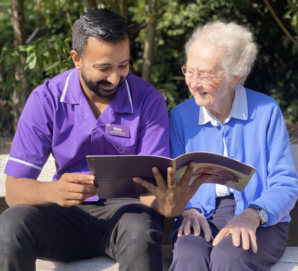 Care home staff member reading with an elderly resident on a bench outdoors, both smiling and engaged with the booklet.