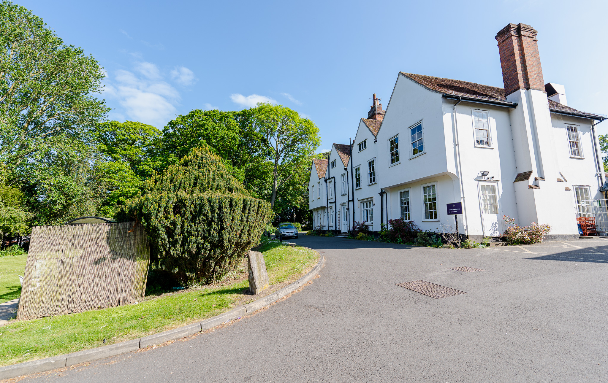 Front view of The New Deanery care home in Braintree, a historic white building surrounded by lush trees and greenery on a bright day.