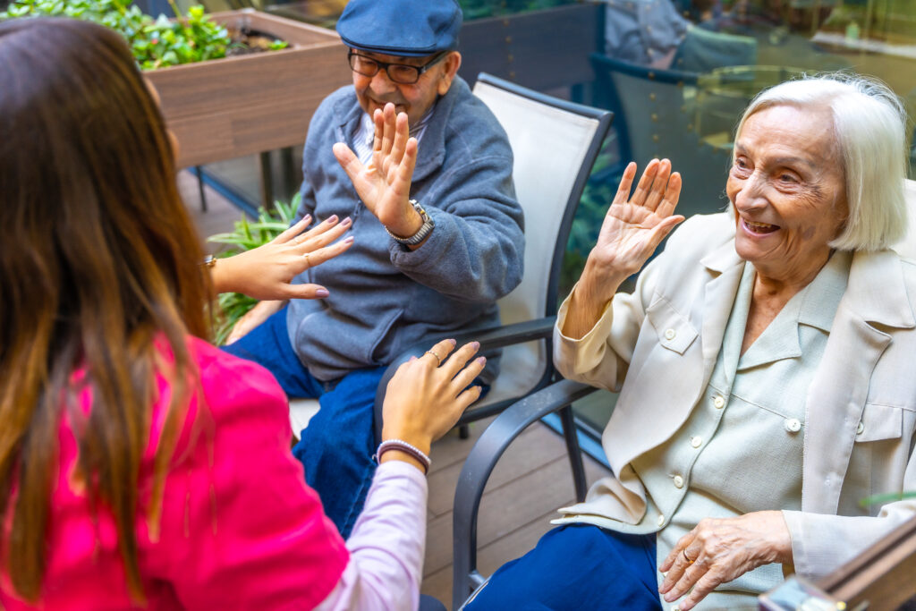 Two elderly people, a man in a cap and a smiling woman with white hair, sit outdoors with a caregiver, enjoying a cheerful interaction.