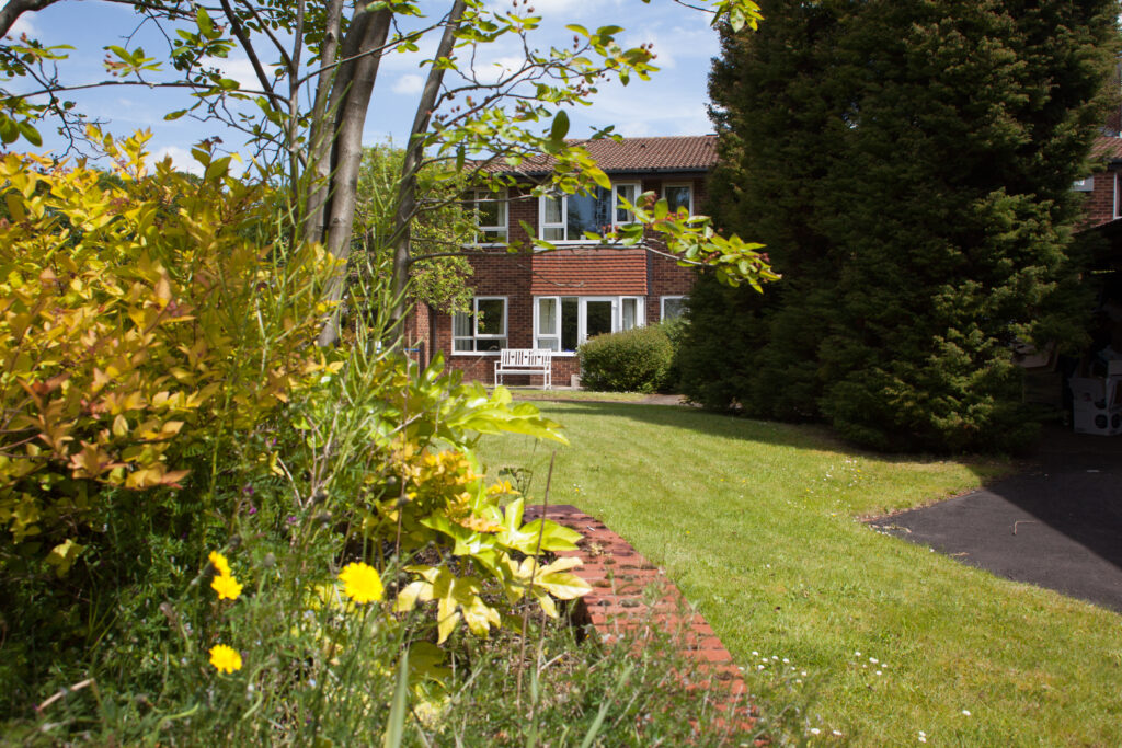 Lush garden with trees and flowers surrounding the exterior of a care home, with a white bench visible in front of the building.