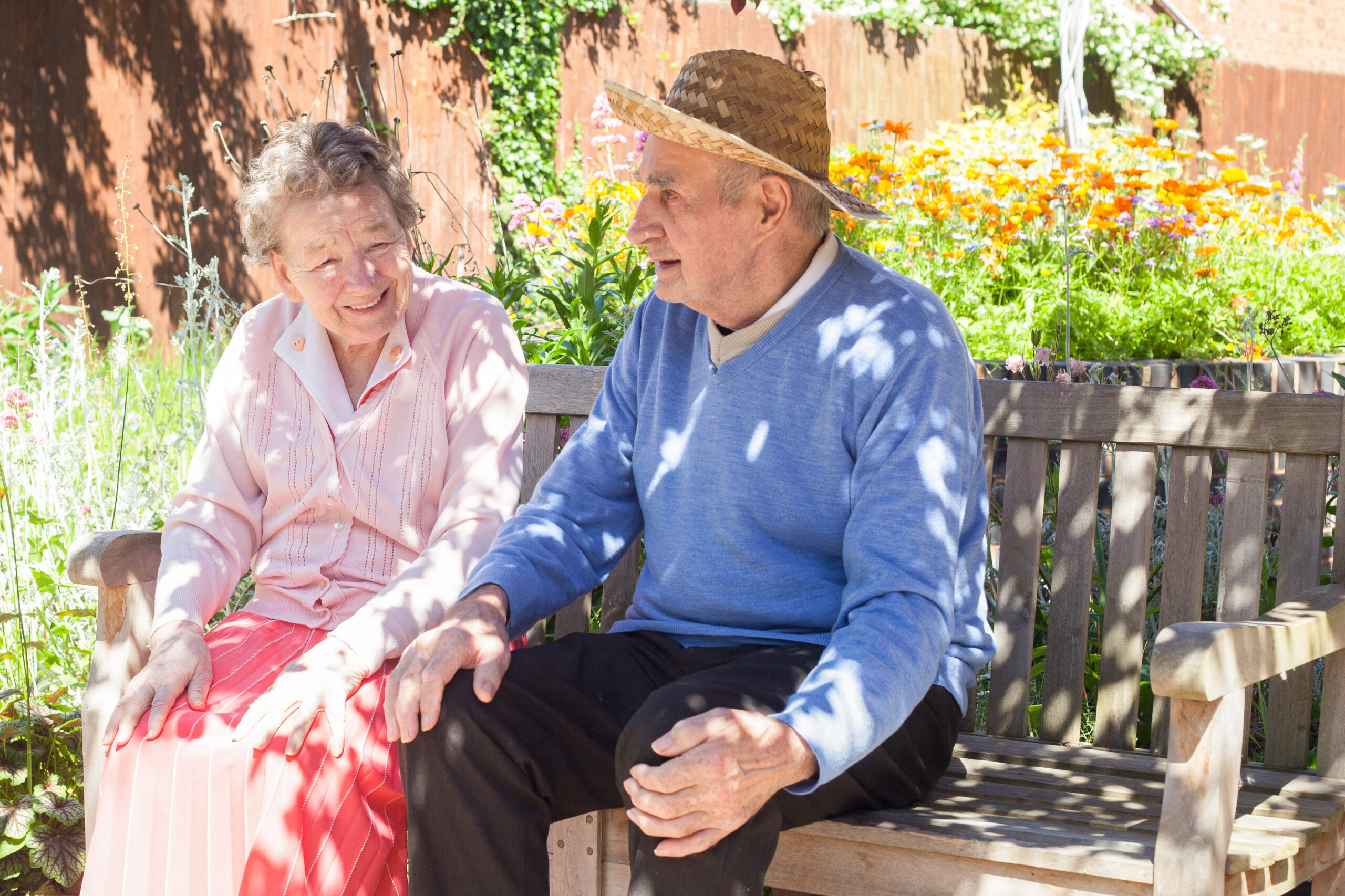 Two elderly residents enjoying a sunny day in the garden at a care home in Ealing, sitting on a wooden bench surrounded by flowers.