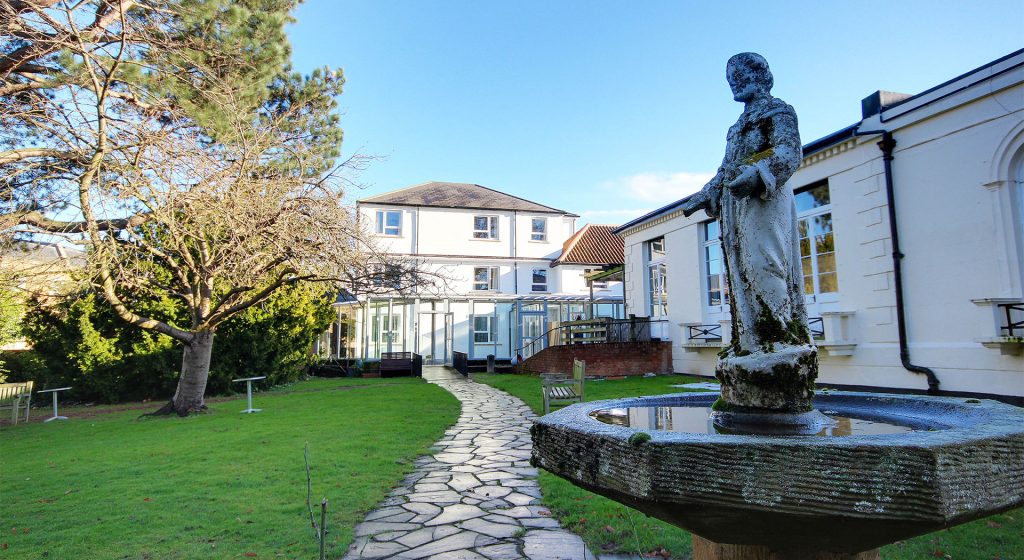 Peaceful garden with a statue and walking path at an Ealing care home, featuring greenery and a classic white building.