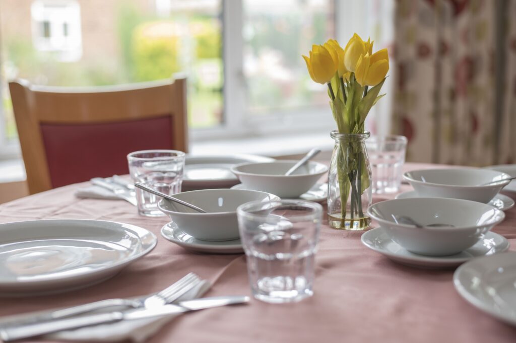 Table set with white dishes and glasses, featuring a small vase of yellow tulips as a centrepiece.