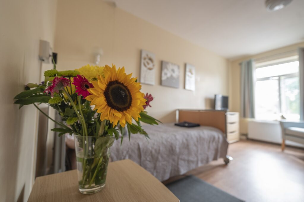 Close-up of a sunflower arrangement in the Martins House care home bedroom with a bed, dresser, and window in the background.
